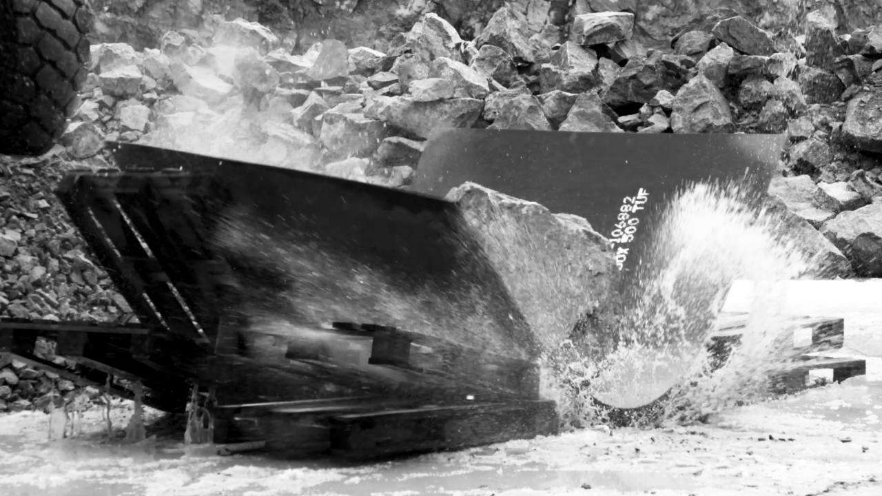 Närbild av ett enormt stenblock som landar på en stålplåt i Hardox® 500 Tuf. Plåten böjs men uppvisar enastående slagtålighet.